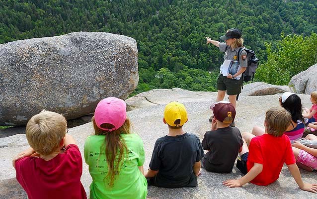 A group of children sitting on a rock overlooking a green valley while a teacher ranger teacher talks