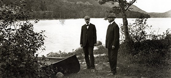 Two men stand near a canoe at the side of a lake. Two men stand near a canoe at the side of a lake.