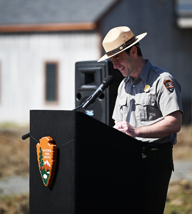 Man in NPS uniform and flat hat stands at a black lecturn with an NPS arrowhead on the front