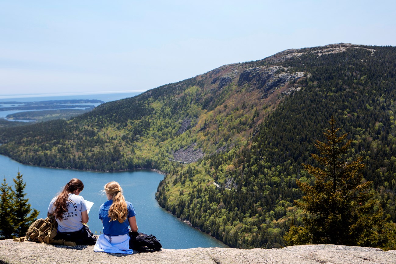 Two people sit on a rock ledge facing a view of water and mountains