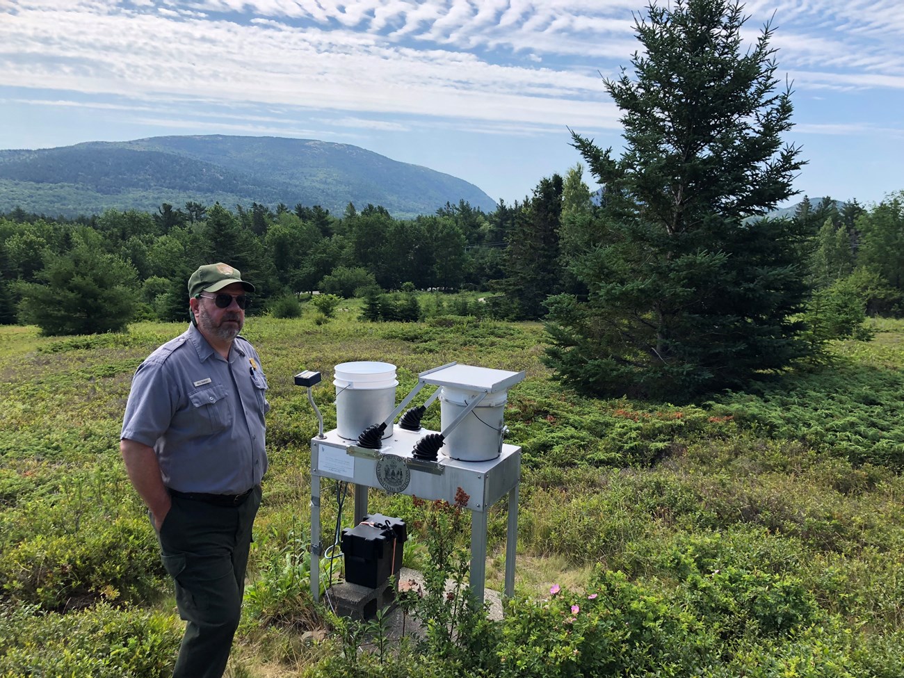 a park ranger standing next to two buckets on a table with weather measuring equipment