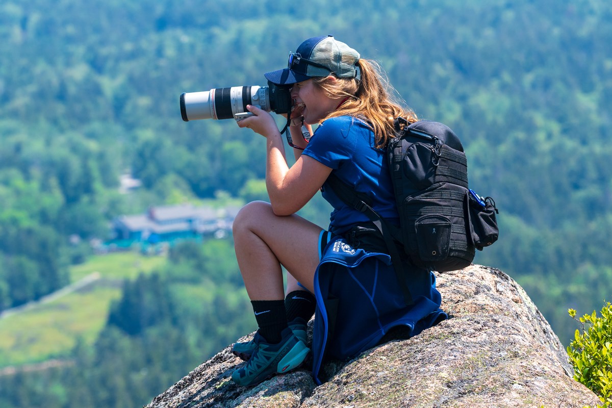 Person crouched on a granite rock, smiling and taking a photo with a large lens.