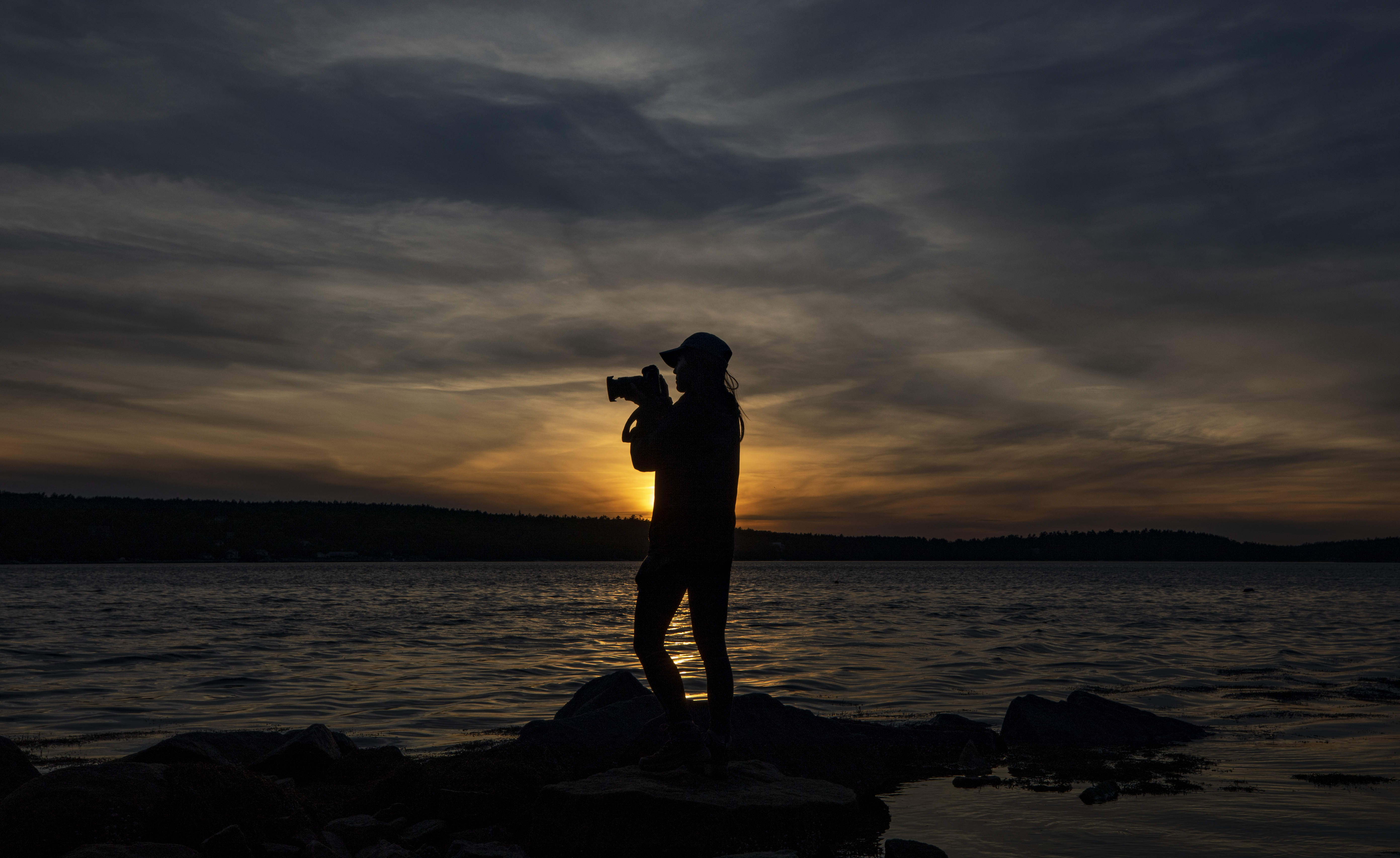 Woman holding camera stands along coastline in front of sunset