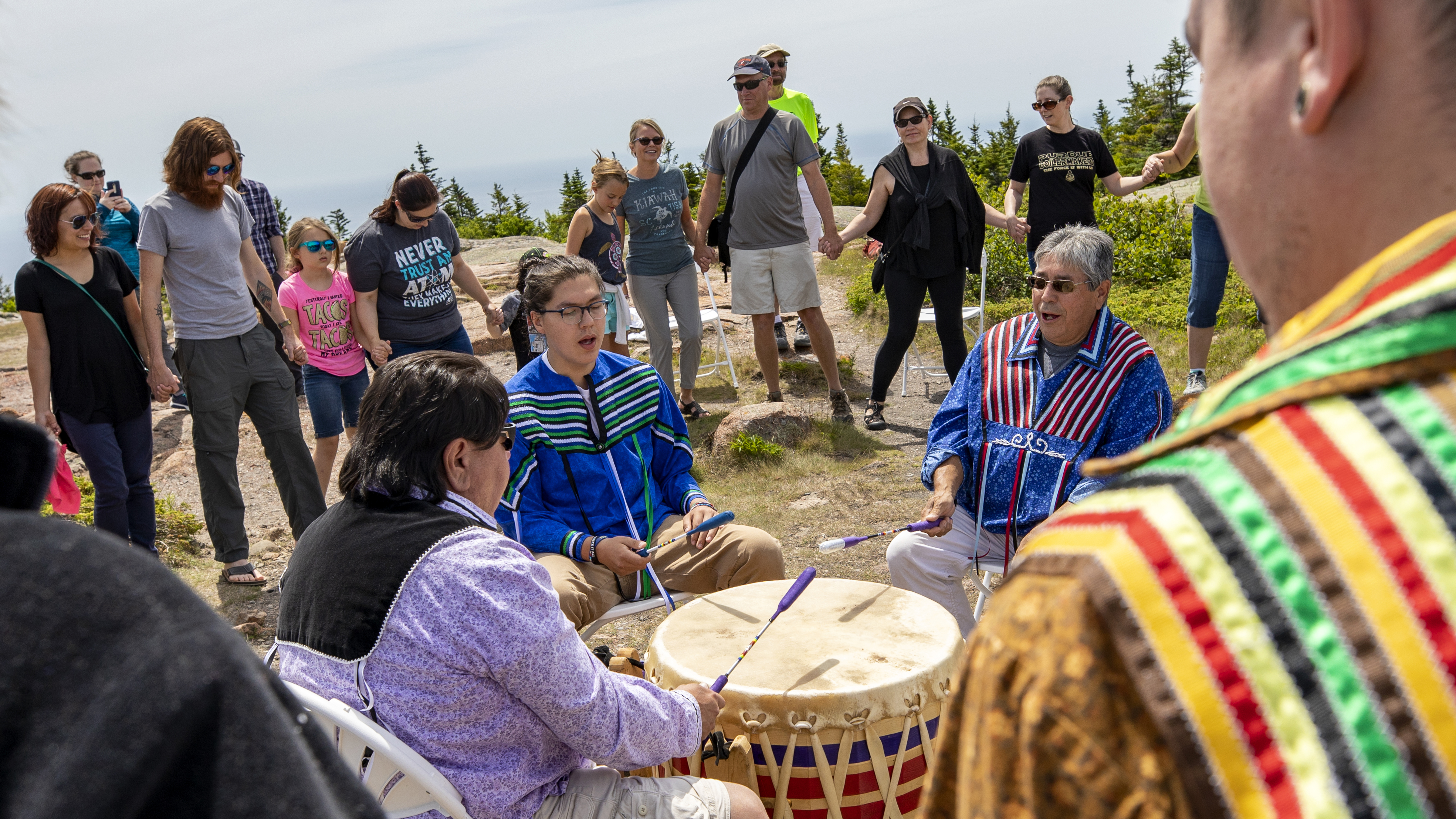 A group of Wabanaki men sit around a drum singing and striking the instrument.