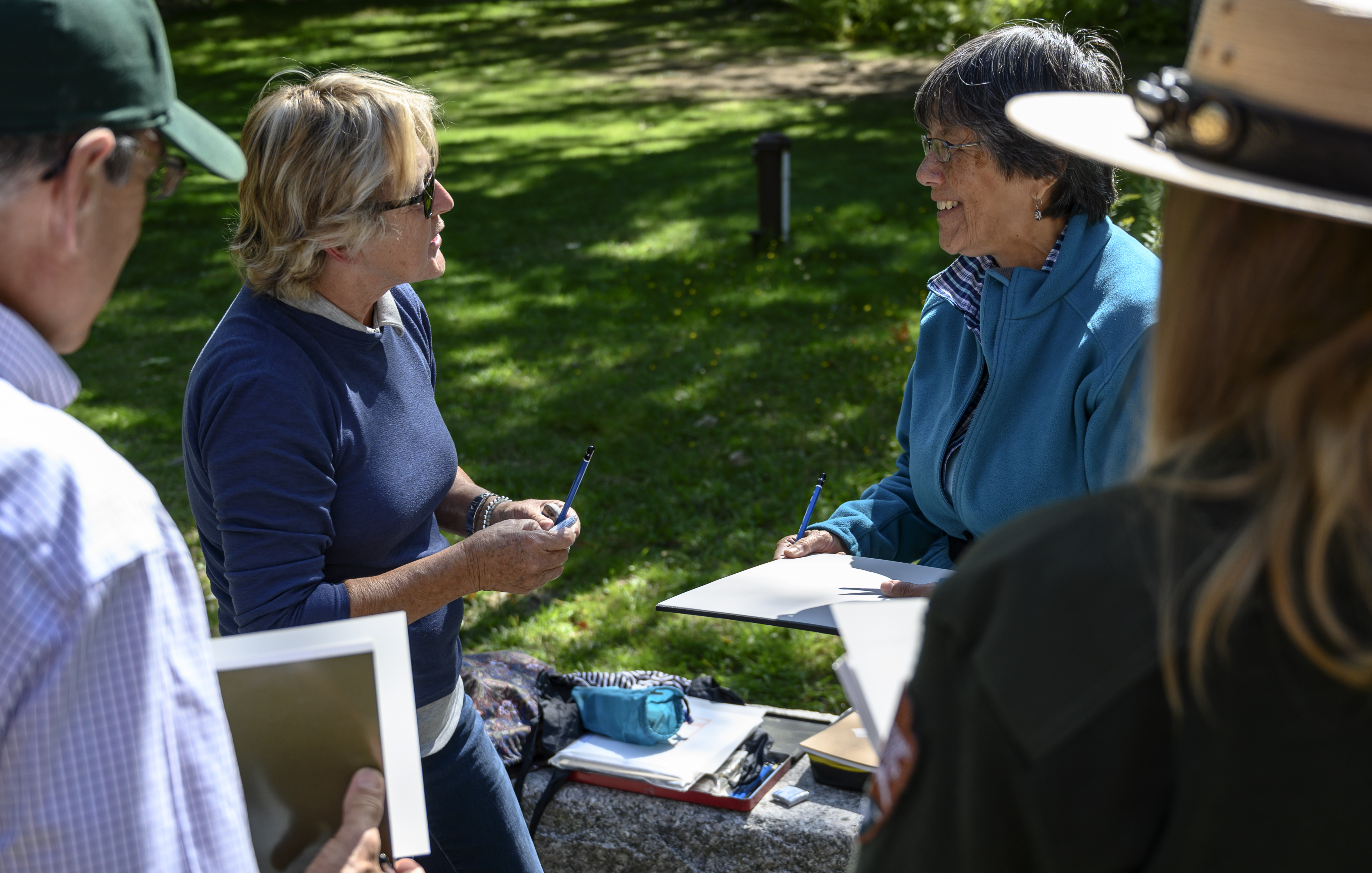 Woman holding drawing pencils speaks with three people, including a ranger in a flat hat, in an open outdoor setting.