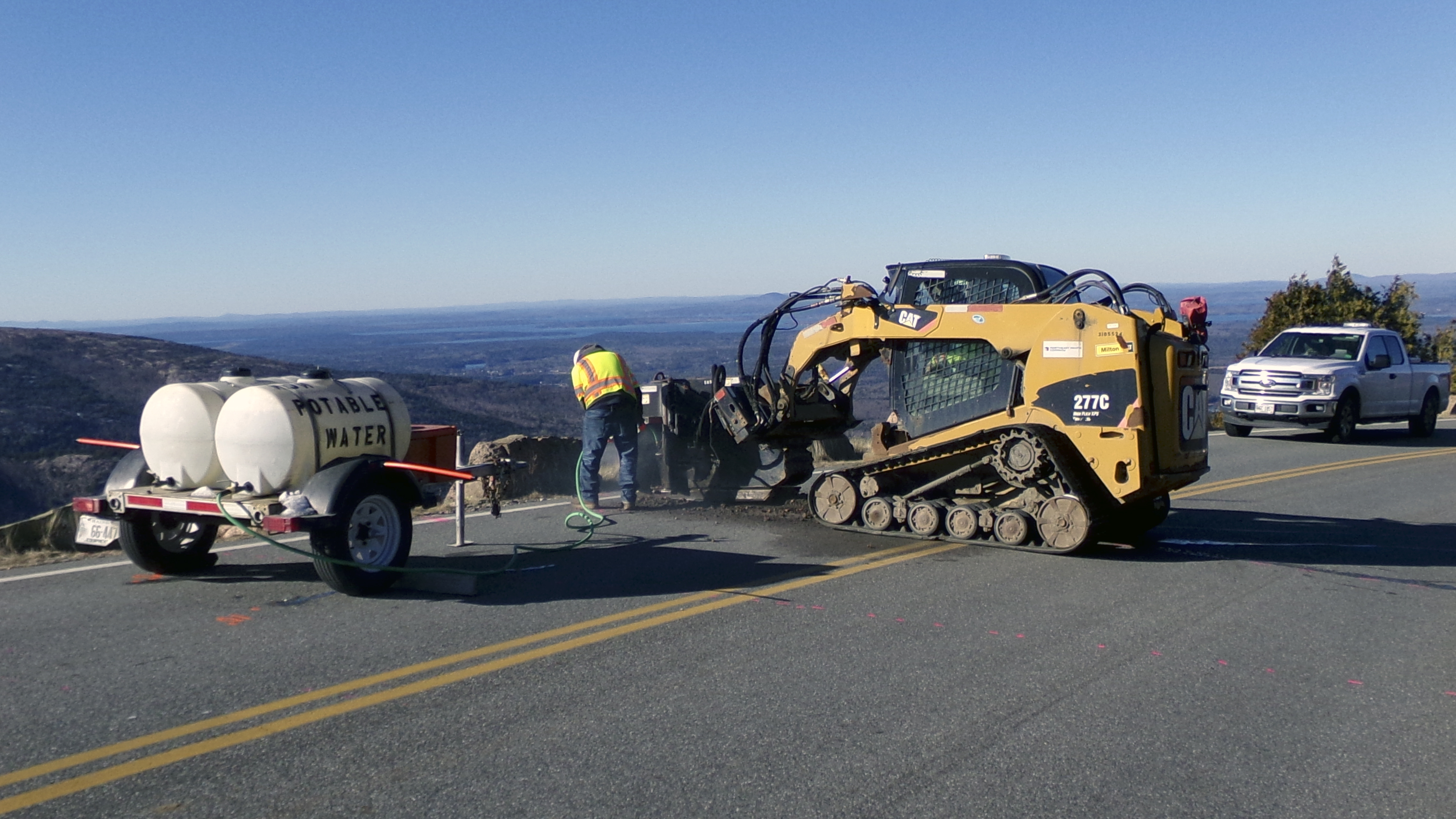 A worker in a hard hat and safety vest stands next to heavy equipment on a mountain roadway A worker in a hard hat and safety vest stands next to heavy equipment on a mountain roadway
