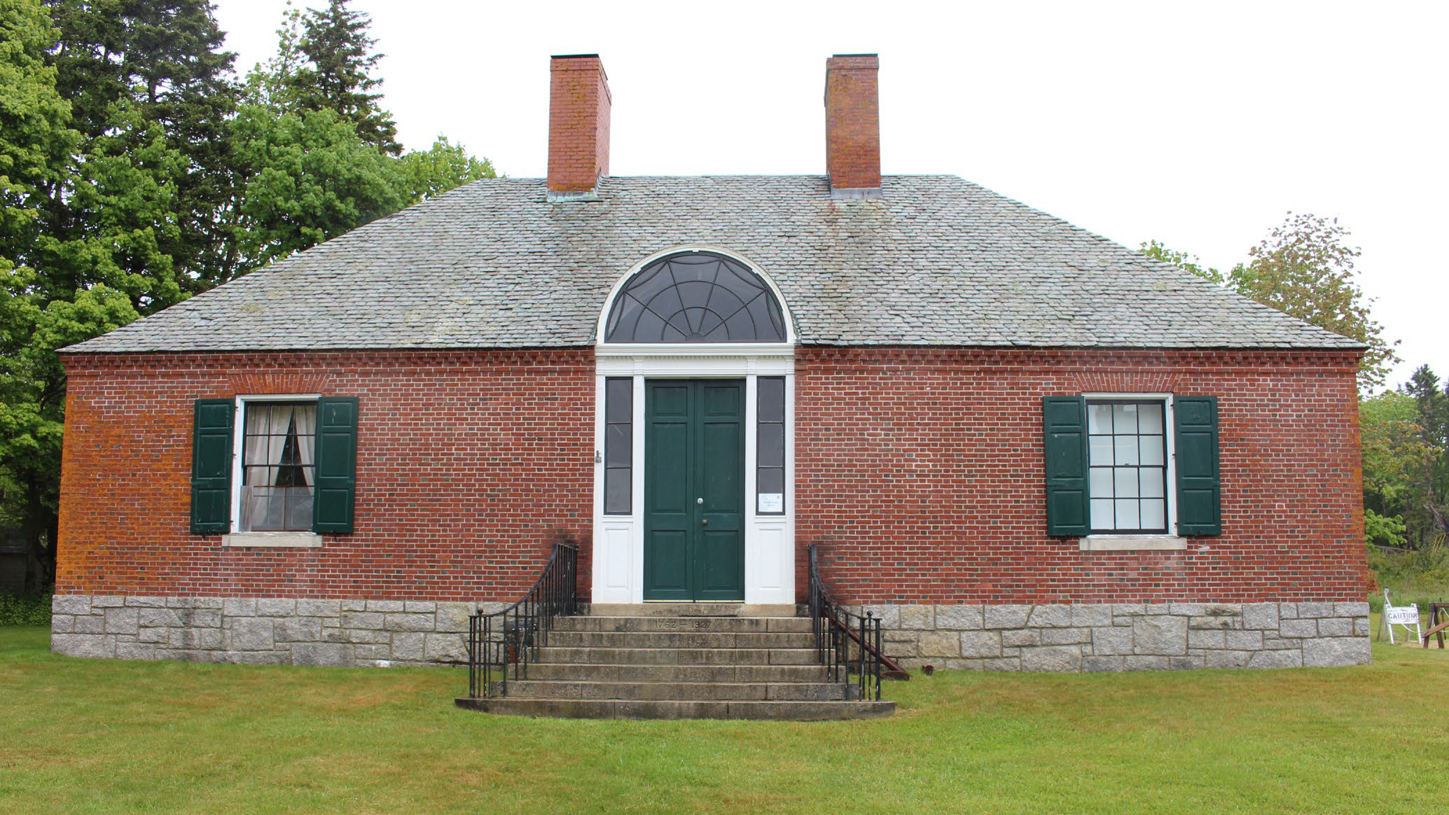 Historic brick building with slate roof, two chimneys, two windows with green shutters, and a green doorway at the center with an arched window above it