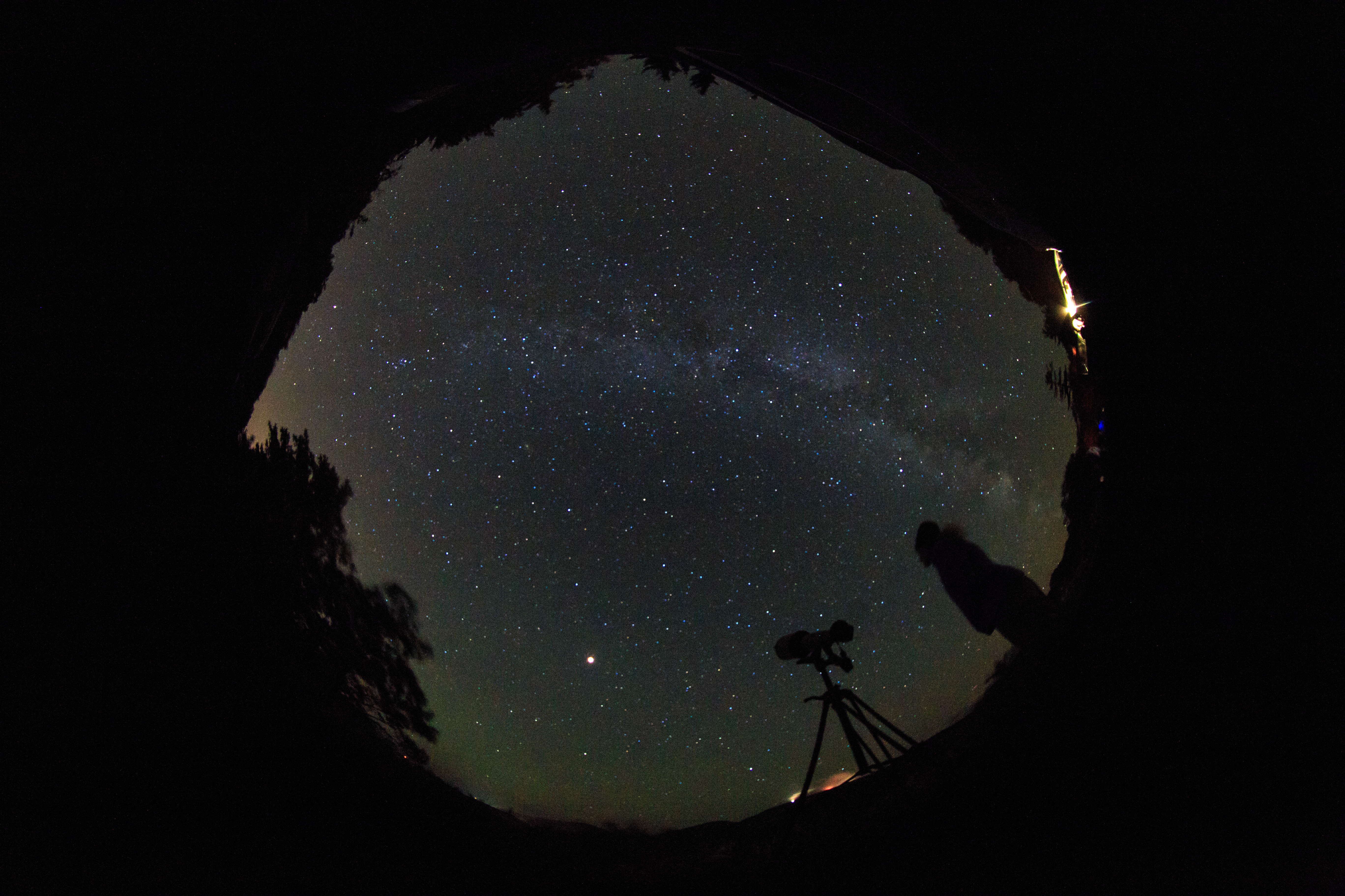 Milky Way with person standing by a long lens on a tripod as seen through a fish-eye lens.