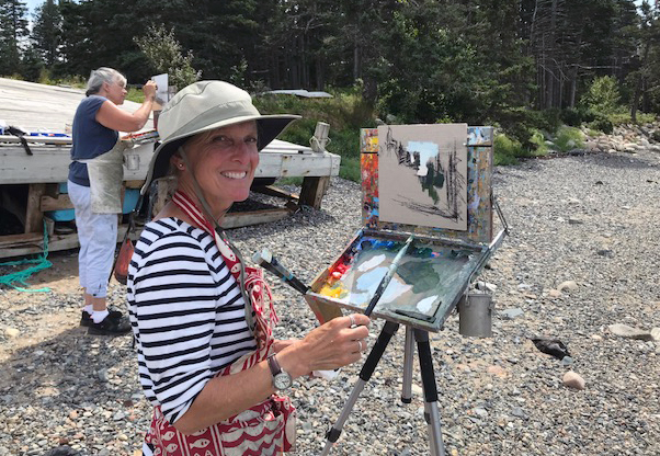 Woman in floppy hat stands by a painting easel along a rocky shoreline