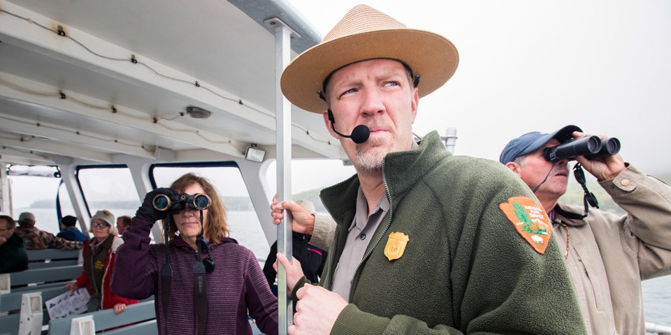 Ranger on boat looks into distance while two visitors look through binoculars Ranger on boat looks into distance while two visitors look through binoculars