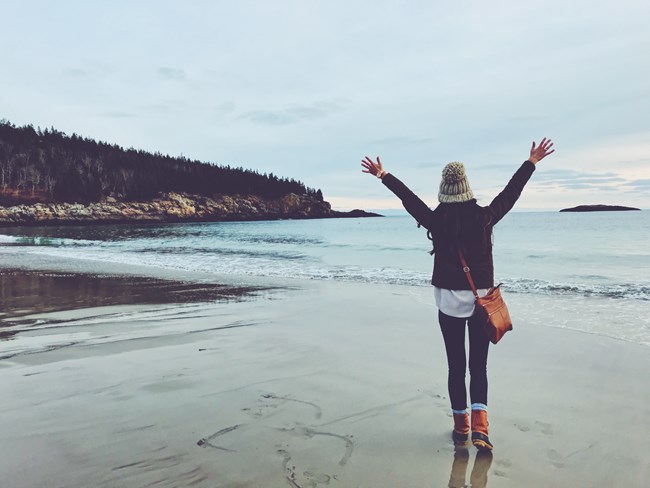 Woman in fluffy knit hat holds her arms outstretched facing the ocean along Sand Beach
