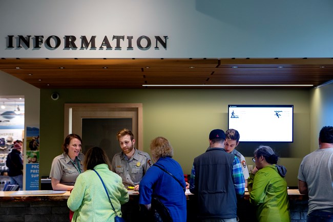 Under a sign saying "information", several rangers talk to a row of people from behind a desk.