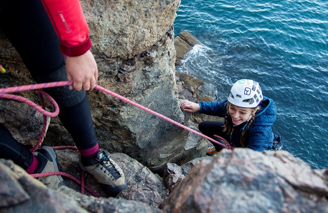 A smiling person in a white helmet climbs a rocky cliff over water. A rope leads from the climber to a person on the left, with only legs and one hand visible.