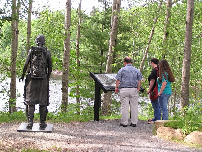 In in outdoor trail setting next to a body of open water, a man and two women look at an interpretive wayside next to a bronze statute of a Native woman