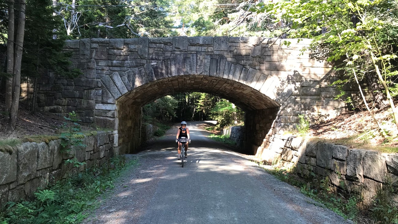 A bicyclist wearing a helmet rides under a carriage road bridge