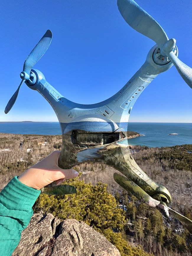 Hand holds a drone painted to mirror surrounding landscape against a view of Atlantic coastline