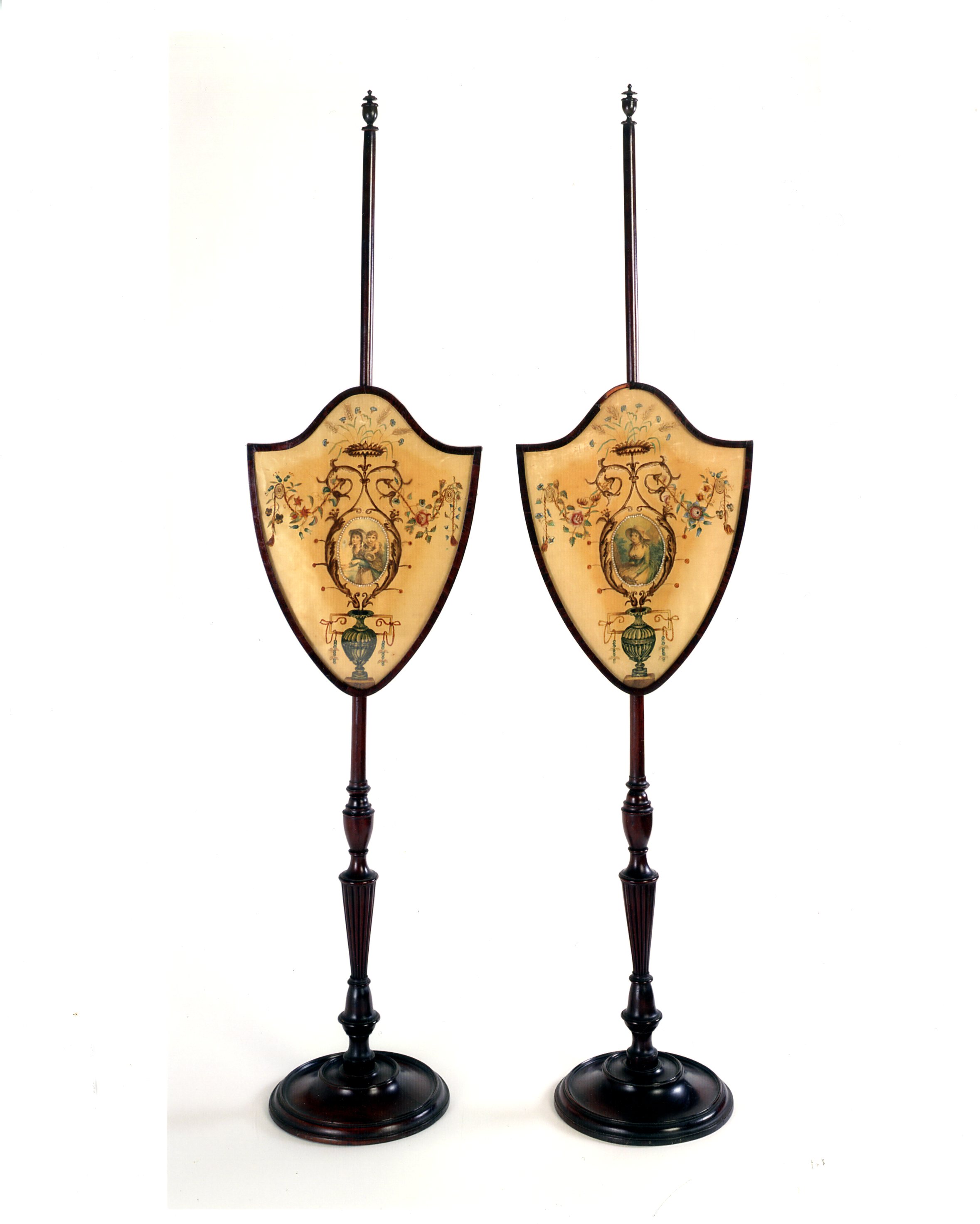 Two pole fire screens stand next to each other on a white background. The screens are identical in shape. They have a round base, an ornamentally curved pole, and screens in the shape of shields. 