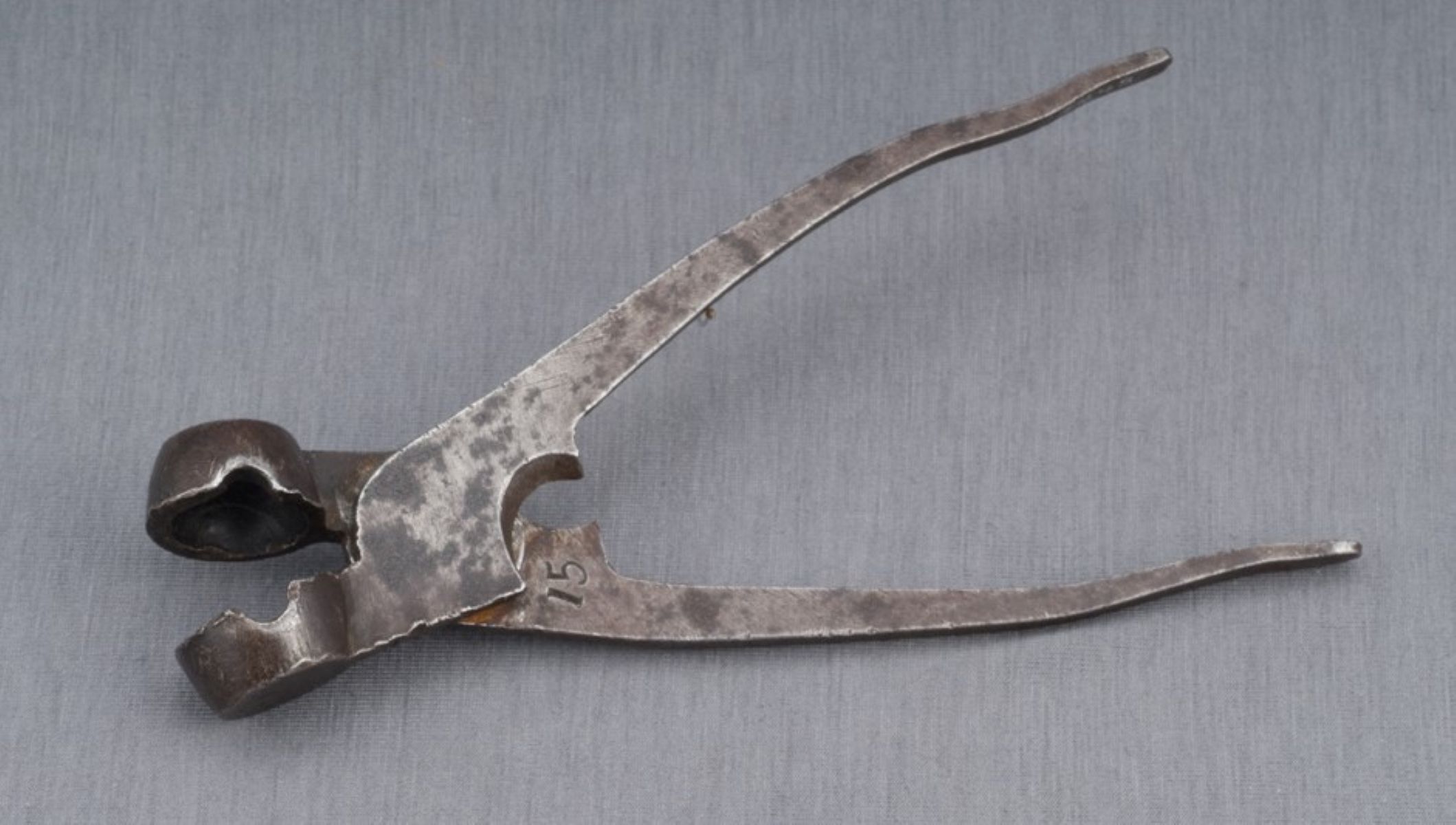 Bullet Mold An iron bullet mold shaped like pliers rests on a grey background. The mold has two semicircle tips that close to form a sphere. The handles are relatively straight and the edges of the mold are damaged, showing signs of use.