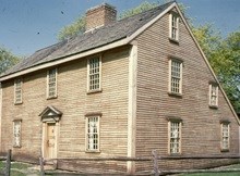 The John Adams Birthplace, a view from the front.  A light brown home with a chimney coming out the center of the roof.  The roof slants dramatically to the back.