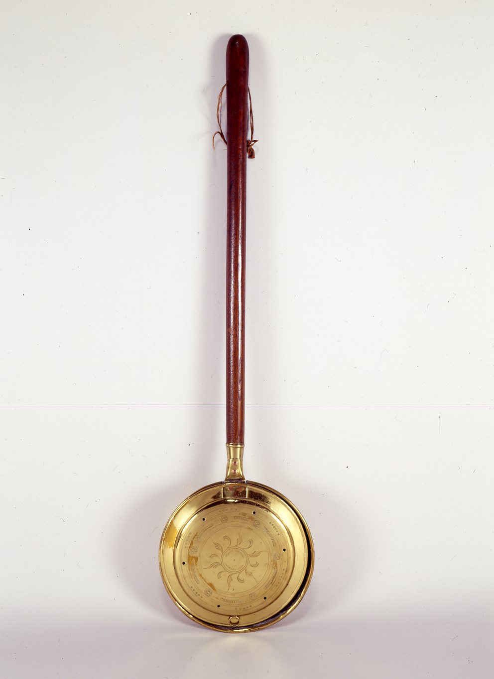 A brass bed warming pan is propped up on a gray background. The circular brass pan with decorative scrolling etchings has an inscription that reads “Abigail Adams, 25th October, 1764.” The pan also features six holes along its perimeter.