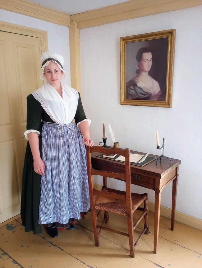 A woman wearing 1700s-style clothing including a long dress, apron, and cap stands next to a historic desk and chair below a portrait of Abigail Adams in a historic room