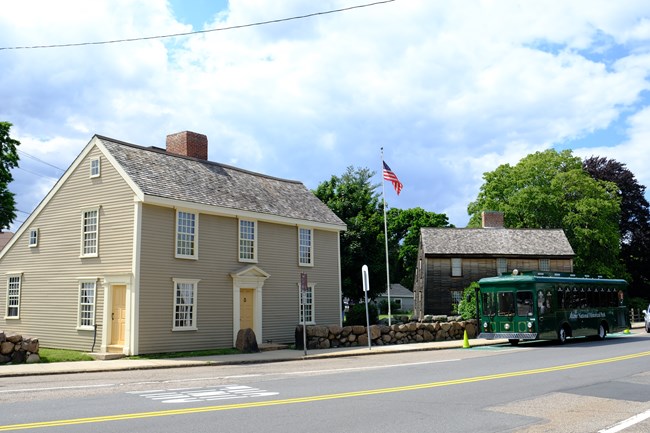 A bus designed to look like a trolley is parking alongside two saltbox-style historic homes while a US flag flies overhead