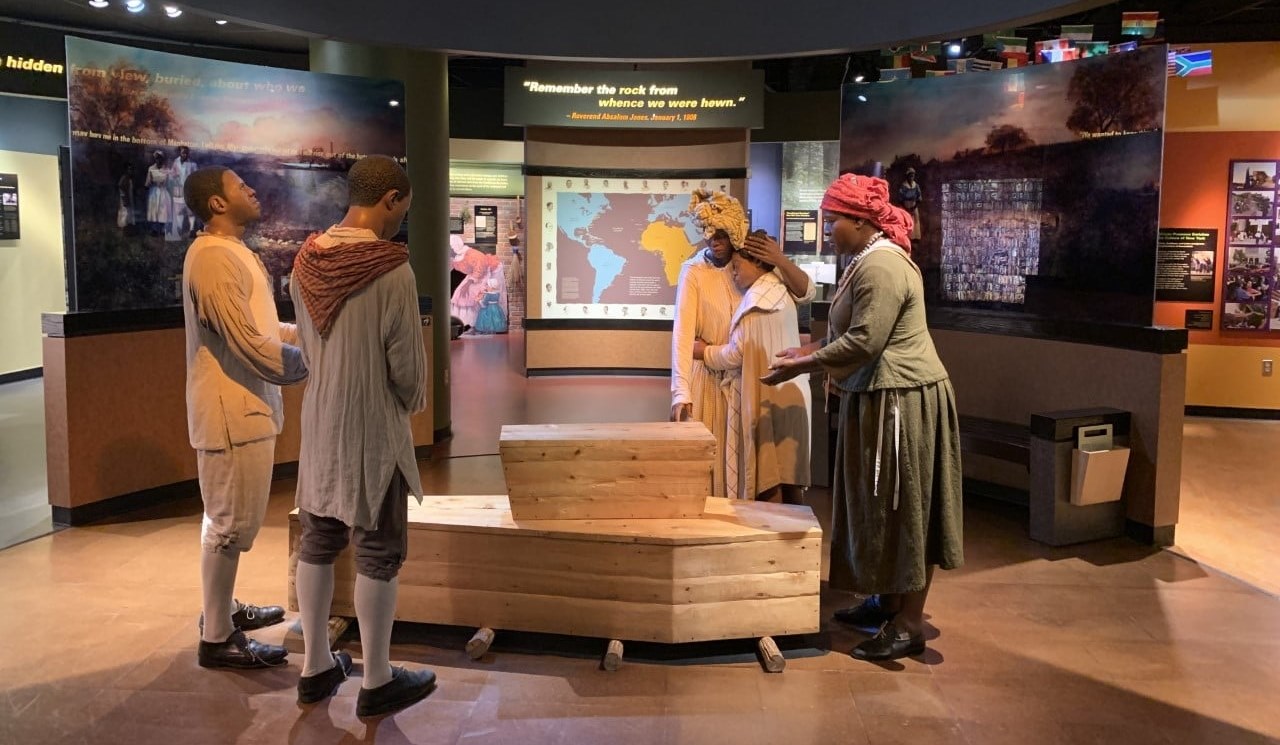 VC Reopening Pic A colored image of the African Burial Ground National Monument’s Visitor Center. Figures depicting a burial scene stand around a small casket sitting atop a large casket.