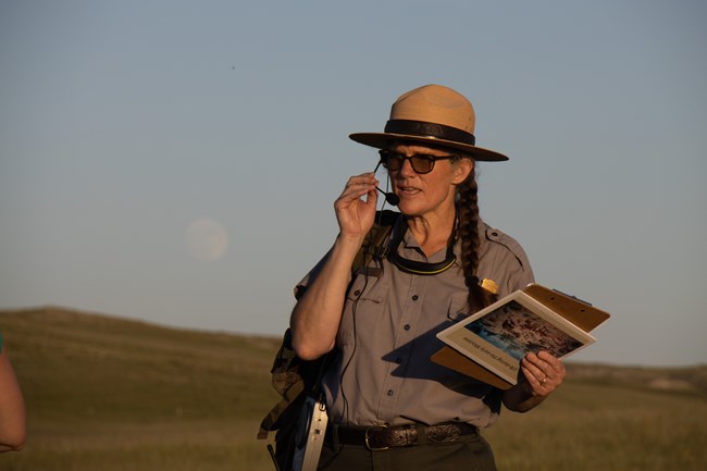 Ranger in uniform talks into a headset microphone while holding a clipboard and pictures outdoors.