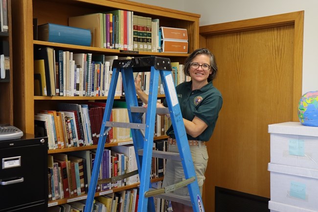 Woman in green volunteer polo stands on a ladder next to a large bookshelf full of books.