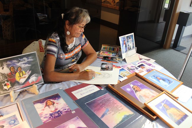 Woman sketches a face from a photo while sitting at a table indoors surrounded by other colored sketches of indigenous people.