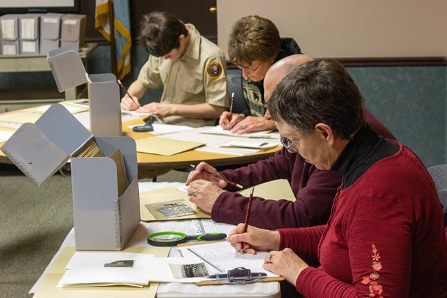 4 people sit behind tables indoors writing with pencils. Magnifying glasses, file boxes, and historic photos lie on the tables.
