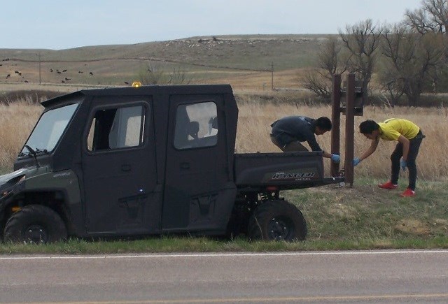 2 people lean over near the posts of a wooden sign. They paint the posts while wearing gloves. A small gator vehicle waits near them.