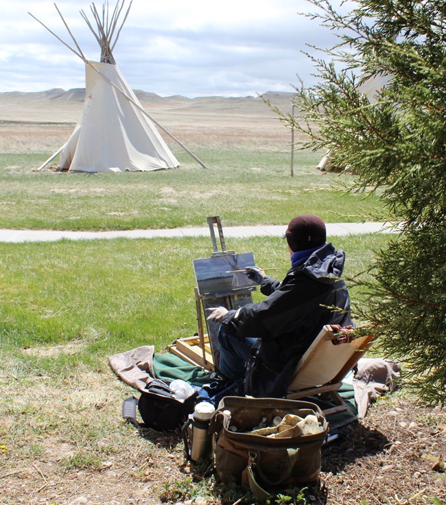 Person sits on the grass in front of a tipi & rolling hills. They paint the same scene on a canvas.