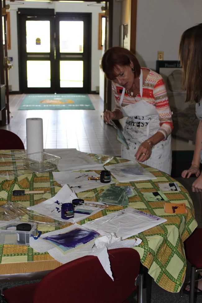 Woman in an apron leans over a table of open paint jars and shiny, painted papers indoors.