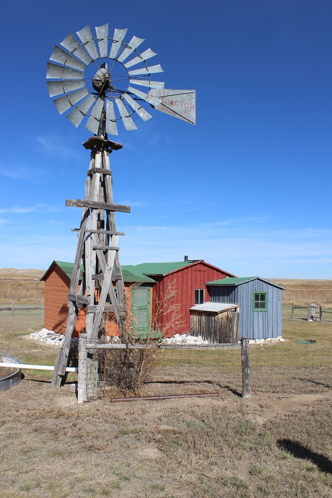 A three-part building with one orange part, one red part and one blue part, stands surrounded by a rock foundation and green prairie with a windmill in the foreground.
