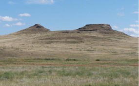 The Fossil Hills stand against a blue sky with a few wispy clouds. Green and brown grassland in the foreground leads to the hills.