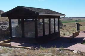 A wood structure with clear sides houses an exhibit on the Daemonelix Trail. A bench and trail marker are outside but face the structure.