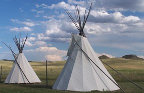 Two triangular shaped, white canvas covered, tipis with gathered poles sticking out of the top stand on a grass covered plain with two fossil hills in the distance and a blue sky with white puffy clouds overhead.