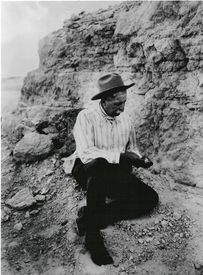 Man in cowboy hat kneels alongside a rock cliff, looking at a stone
