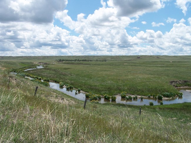 A 4 foot wide spring-fed river carves a giant U in the prairie landscape. There are small clumps of vegetation on the narrow banks of the river.