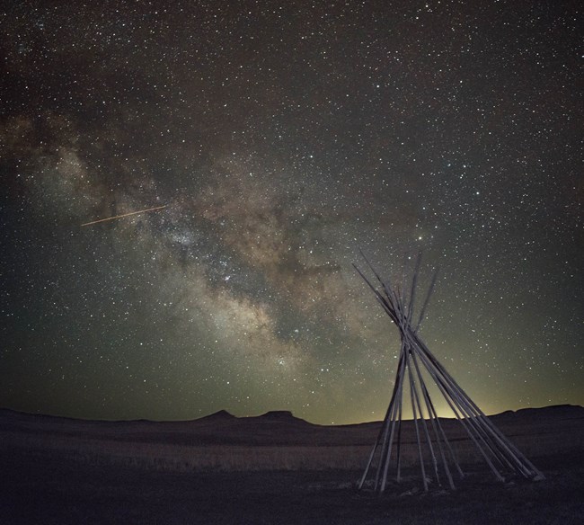 Starry night sky and band of the milky way galaxy above silhouetted hills and soft lit tipi poles.