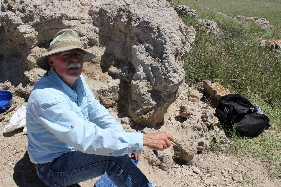 Dr Robert Hunt discuses fossil finds at Agate Fossil Beds Man in hat crouching near a rock formation