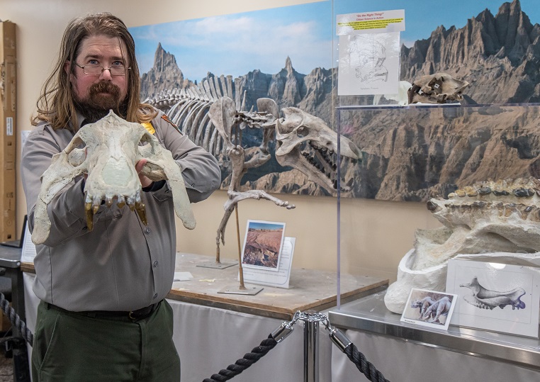 Ranger Ed Welsh A ranger in uniform with shoulder length brown hair, mustache, goatee, and wire-rimmed glasses holds an animal skeleton's skull while standing indoors in front of a full model skeleton.