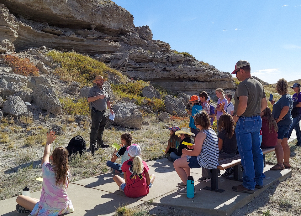 Fall Science Day2021_TeraLynnGray-4 A ranger in uniform stands in front of a hill while talking to about 30 kids and adults. The rocky hill has a rugged, concave section removed from it.