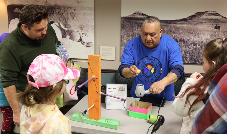 Man in blue OLC shirt holds a ping pong ball over an upturned hairdryer. A wooden post on the table in front of him has perpendicular sticks with rings on each end. A child & an adult watch.