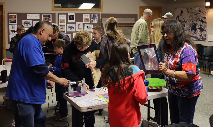 2 adults work with kids around a table of 3 science items. Other visitors explore in the background.