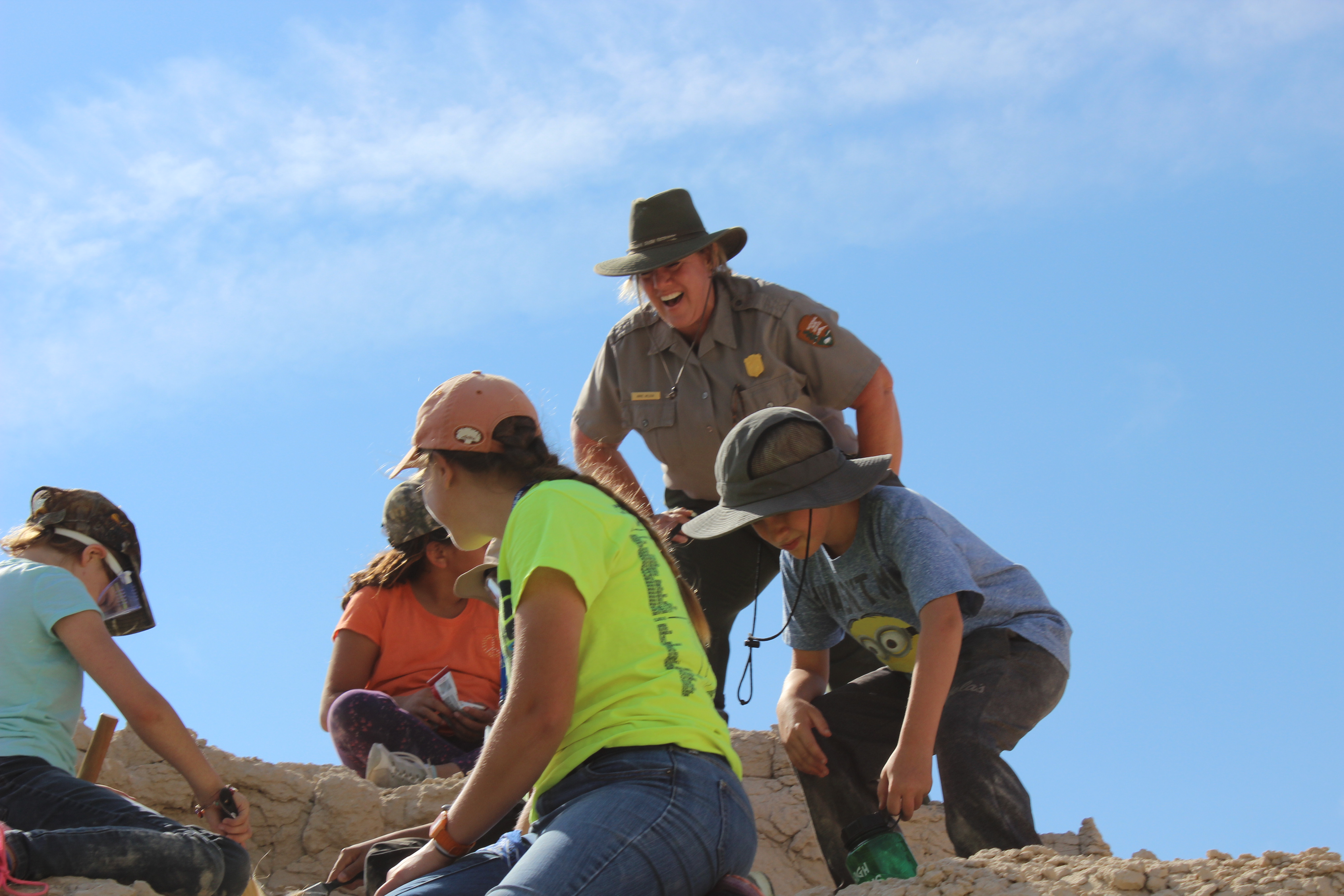 A smiling blond ranger in uniform and hat crouches over observing 4 kids wearing bright clothes and hats excavating fossils on a hill of dirt under a clear blue sky.