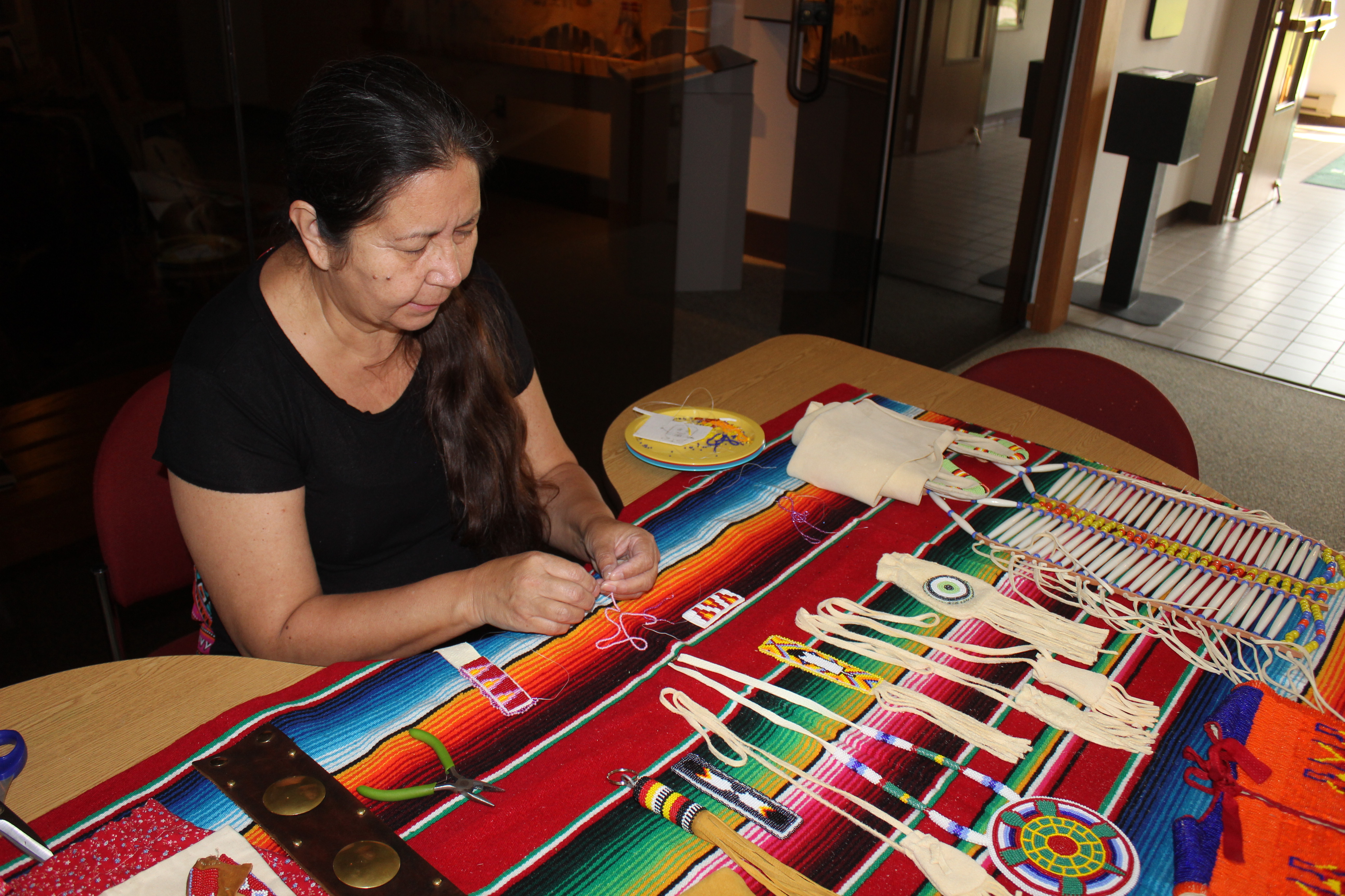 Woman holds string in between her fingers of both hands that rest on a brightly colored cloth indoors. Many small items made with beads are displayed on the cloth.