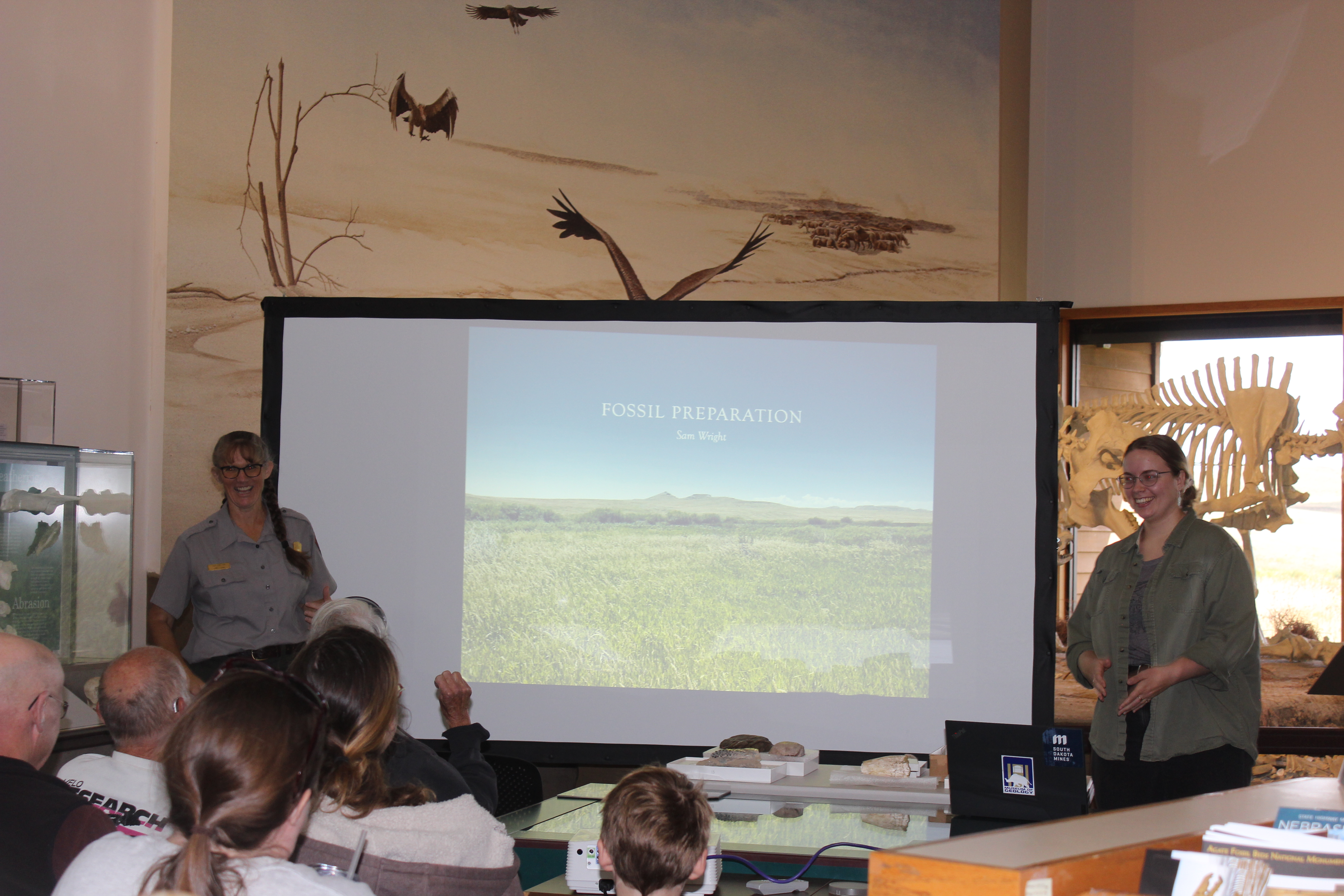 2 women stand on either side of a projector screen. 1 wears an NPS uniform. Several people sit in the audience.