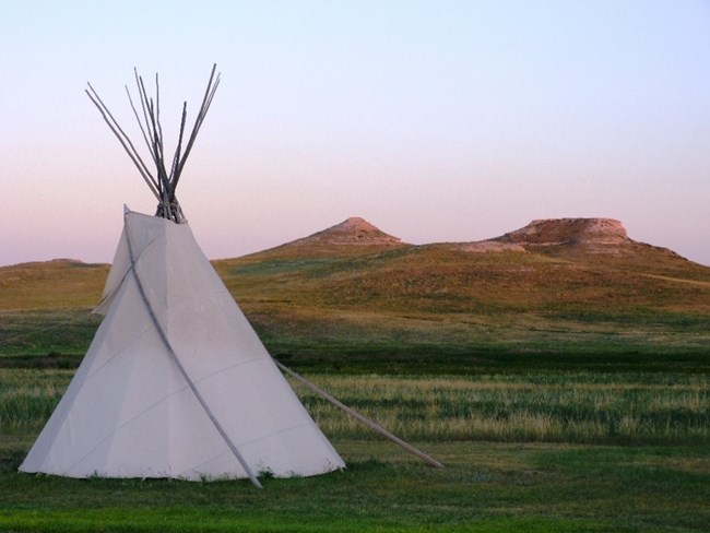 A white tipi stands on green grass in front of sunset lit hills at a distance in the background. The sky is clear blue tinged with purple and pink.